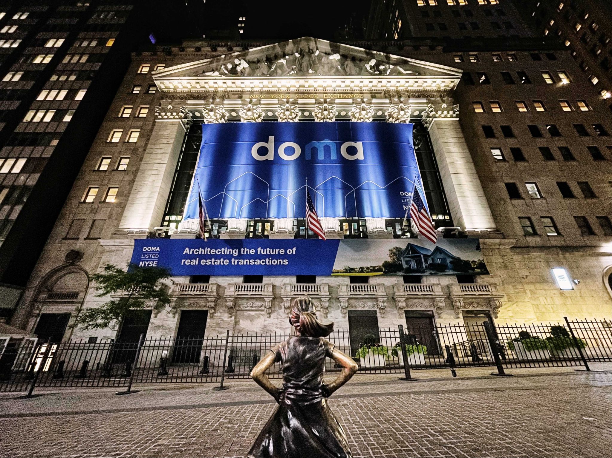 The New York Stock Exchange at night, lit up with the Doma banners, the Fearless Girl statue in the foreground